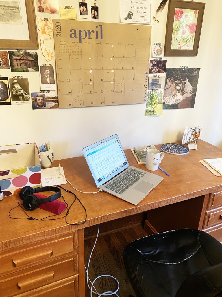 A photo of a desk with a laptop, coffee mug, calendar, and various images and office supplies to illustrate a work-from-home space.