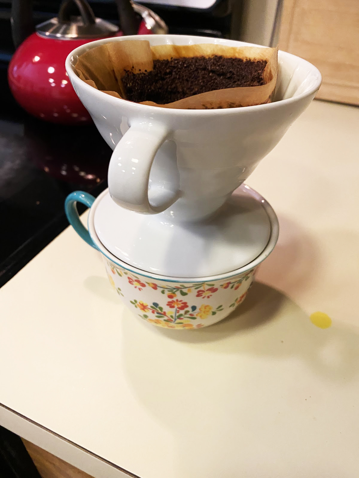 A photo of a single-cup pour over coffee brewer sitting on top of a floral print coffee cup on a counter with a red kettle in the background.