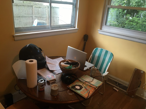 A photo of a messy brown table in a yellow room with candles, paper towels, books, cards, a bowl of bric-a-brac, a motorcycle helmet, and other items cluttering it.