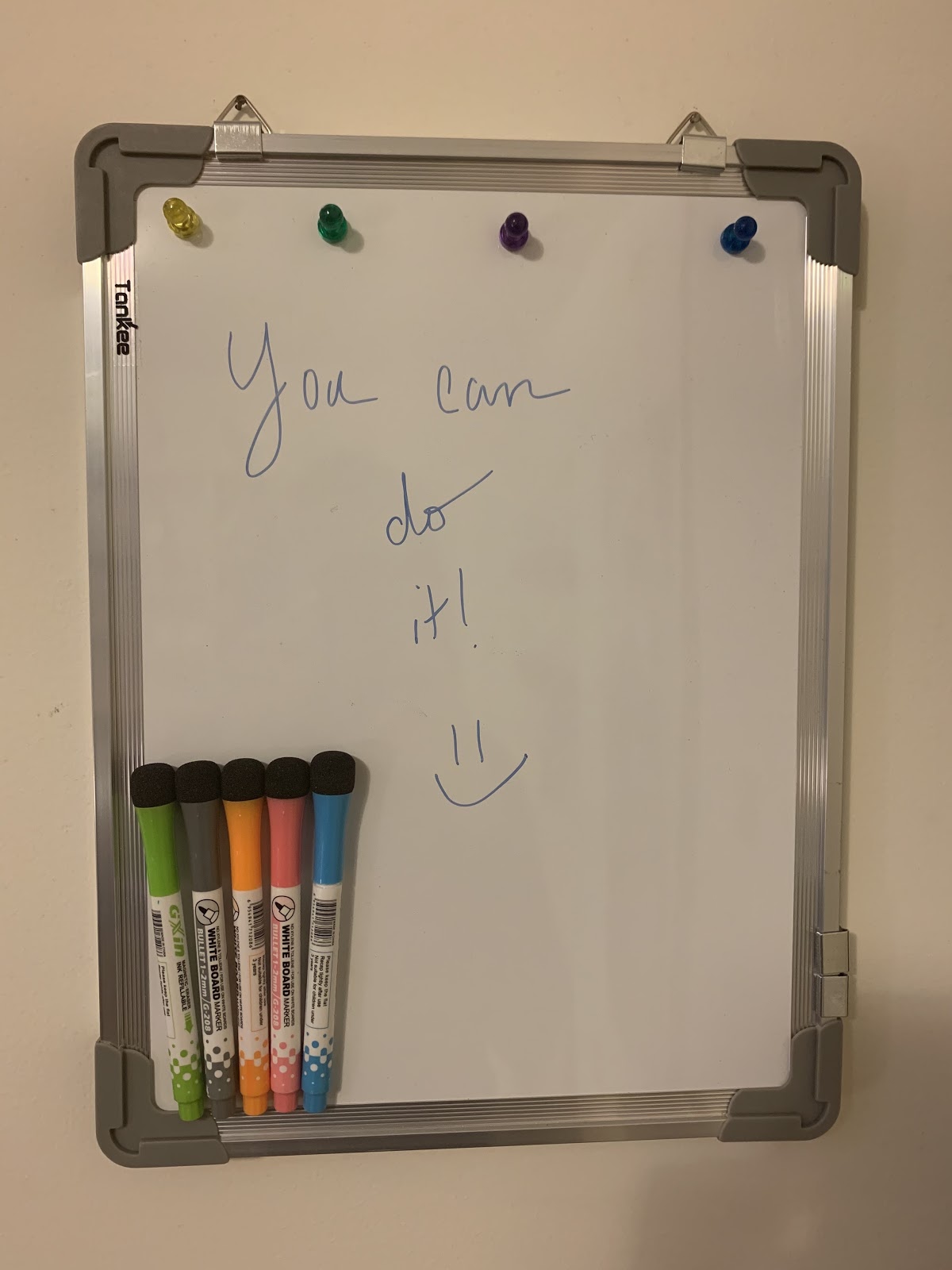 A whiteboard with a message hangs on a wall with five colored markers. The message says "You can do it!" with a smiley face.