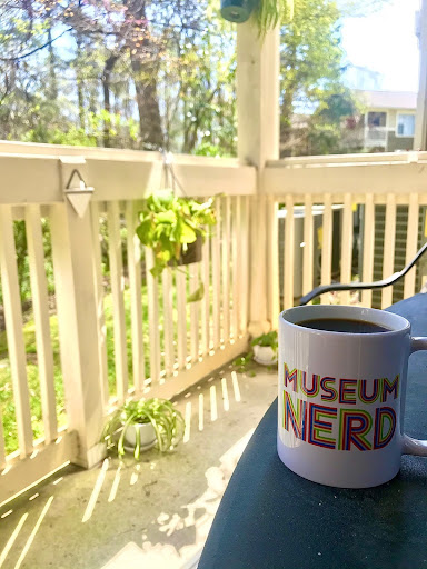 A cup of coffee inside a mug reading "Museum Nerd." The cup is sitting on a small table on my patio overlooking a small wooded area.