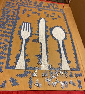 The puzzle in progress. Completed are three white utensils (a fork, knife, and spoon) in the center of the puzzle with an entirely blue border of completed pieces. 
