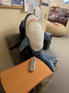 Sharky, the UNC Writing and Learning center mascot, sitting and relaxing in a massage chair. Sharky is a large stuffed shark that enjoys his time at the center!