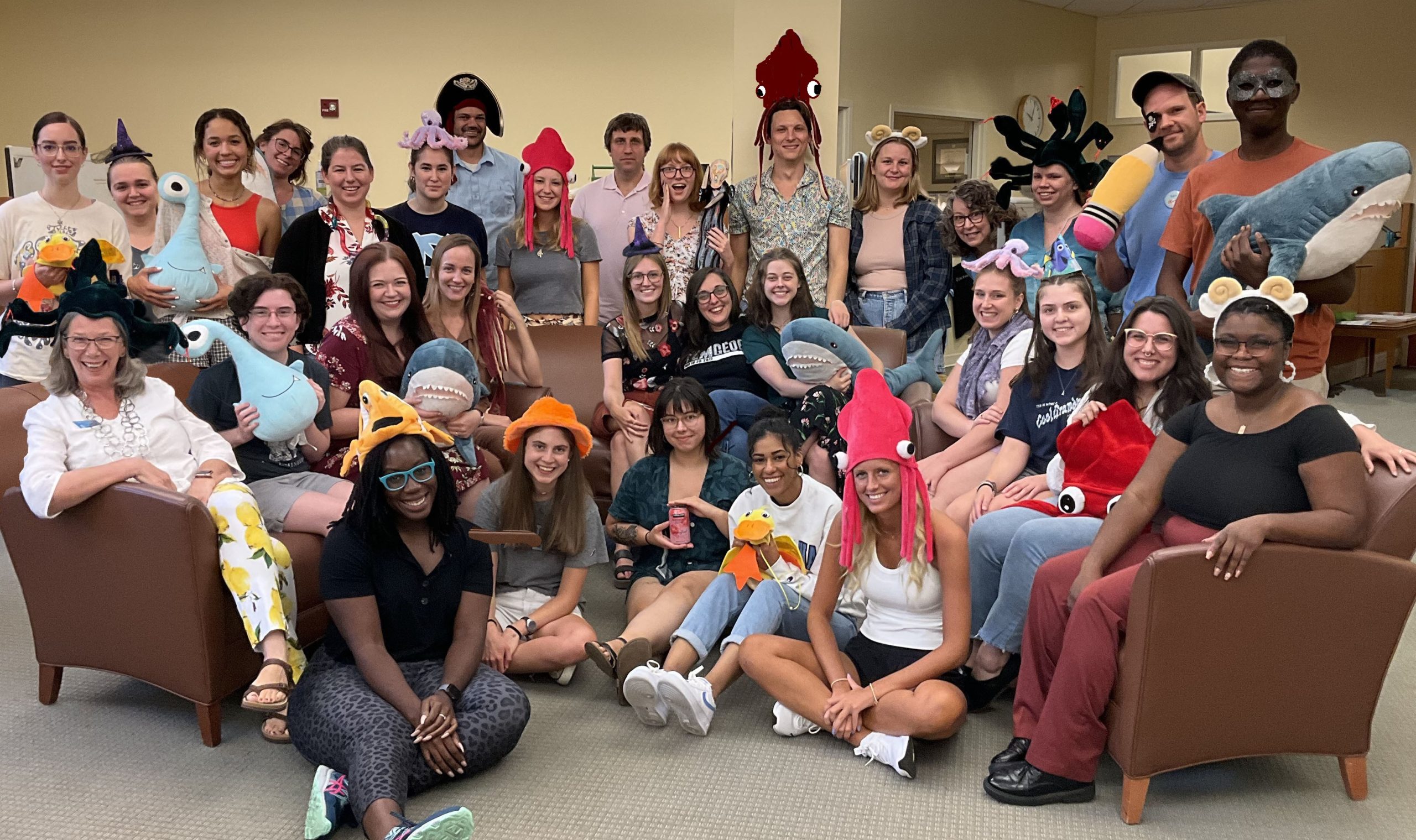 A photo of writing center staff posing together in a group photo. There are about 30 people clustered together over two couches, with some people standing behind the couches and a few people sitting on the floor. Many people are wearing silly hats or holding stuffed animal toys.