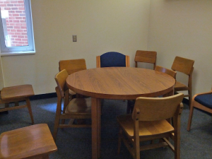 Study Room in Davis Library. A beige room with a corner window. Inside the room is one circular, wooden table with four chair. Four more chairs are show around the room. 