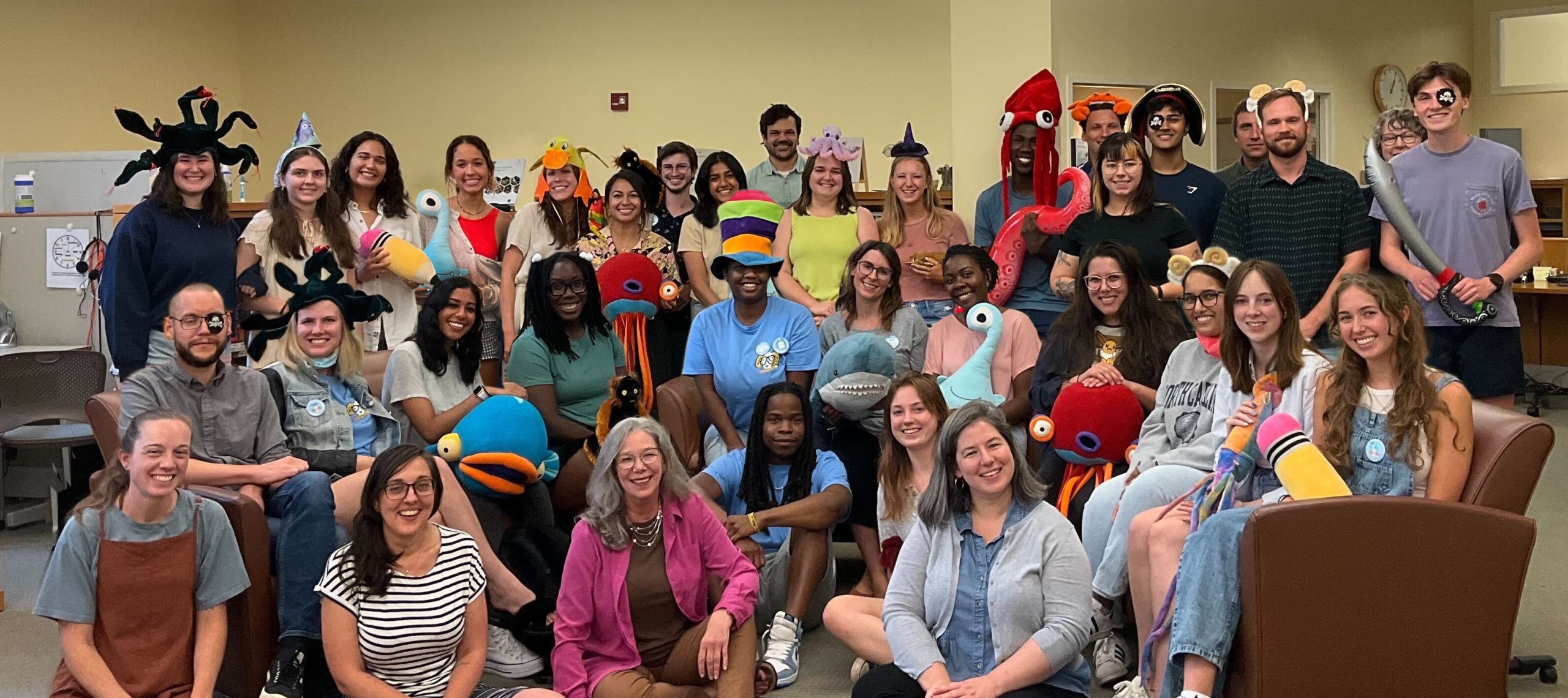 A photo of writing center staff posing together in a group photo. There are about 30 people clustered together over two couches, with some people standing behind the couches and a few people sitting on the floor. Many people are wearing silly hats or holding stuffed animal toys.